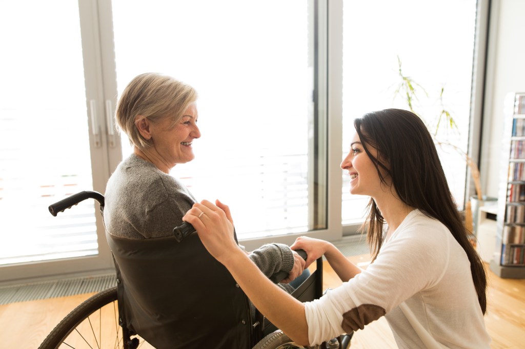 Disabled senior woman in wheelchair at home in her living room, with her young daughter caring for her.